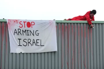 Aκτιβιστής με κόκκινα ρούχα βρίσκεται σε μια στέγη δίπλα σε ένα λευκό πανό που γράφει «σταματήστε να εξοπλίζετε το Ισραήλ».