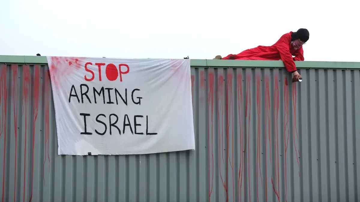 Aκτιβιστής με κόκκινα ρούχα βρίσκεται σε μια στέγη δίπλα σε ένα λευκό πανό που γράφει «σταματήστε να εξοπλίζετε το Ισραήλ».